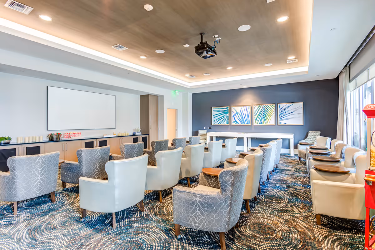 A modern meeting or presentation room with rows of upholstered chairs facing a large white projection screen. The room features a patterned carpet, a wooden ceiling with recessed lighting, and framed abstract leaf artwork on a dark accent wall. There is a popcorn machine visible on the right side near large windows with curtains.