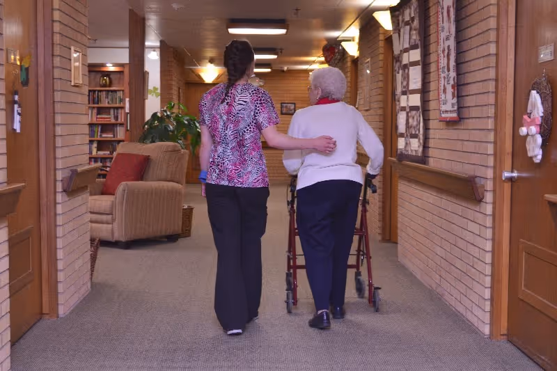A caregiver walking down a hallway with an elderly woman using a walker in a senior living facility. The hallway has brick walls, handrails, and some framed decorations. There is a comfortable chair and a bookshelf visible in a side area.