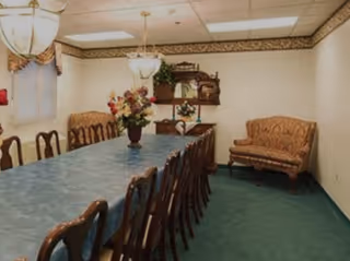 A formal dining room with a long table covered by a blue tablecloth surrounded by wooden chairs. There are two chandeliers hanging from the ceiling, a floral centerpiece on the table, a patterned loveseat against the wall, and a wooden hutch with decorative items in the background. The room has green carpet and cream-colored walls with a decorative border near the ceiling.