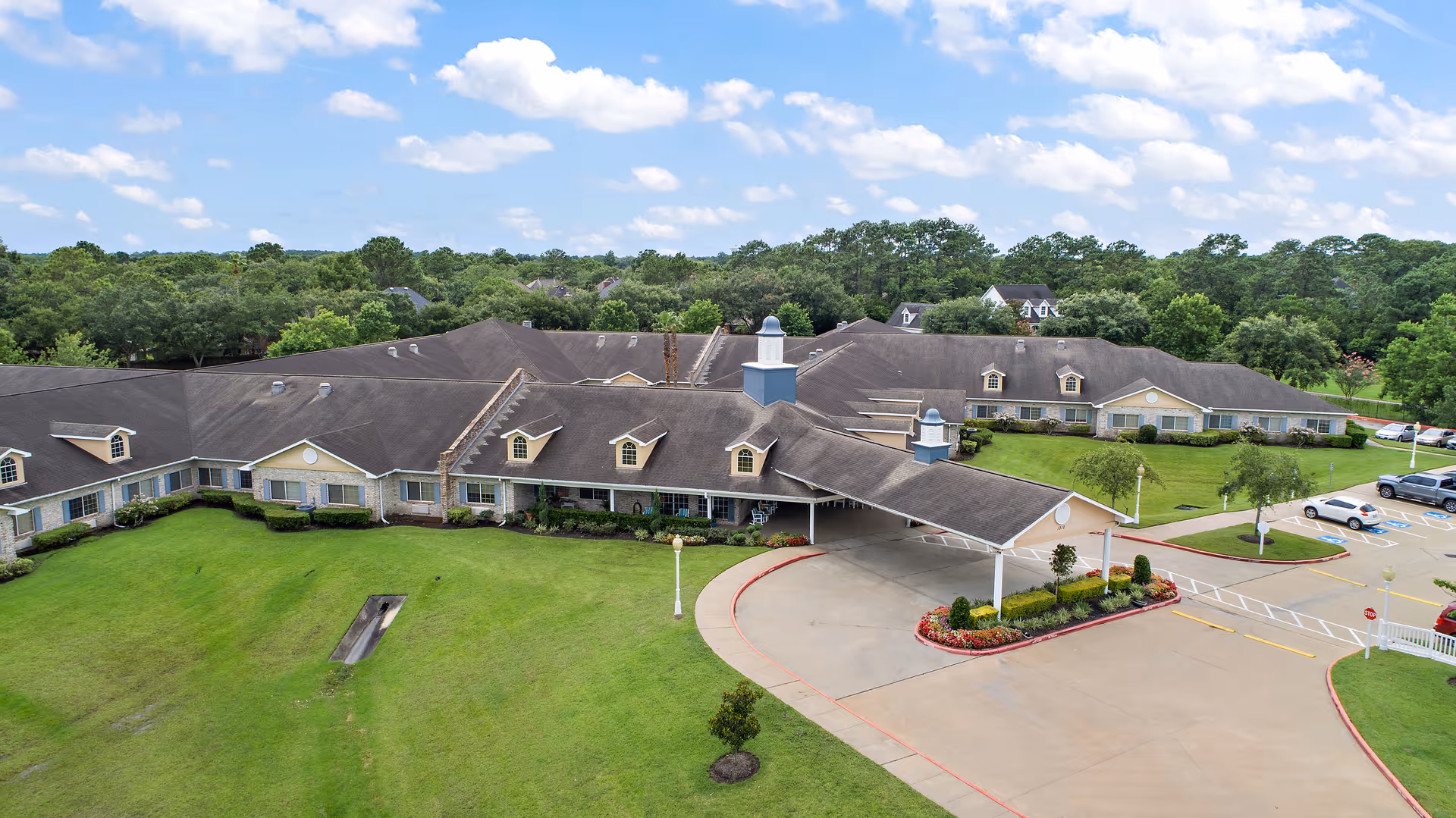 Aerial view of Emeritus at Friendswood senior living facility showing a large single-story building with multiple roof peaks, surrounded by green lawns and trees under a partly cloudy sky. The entrance has a covered driveway with landscaped flower beds and a parking area with several cars.