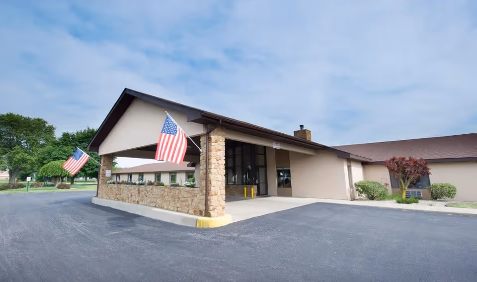 Exterior view of Wesleyan Health and Rehabilitation Center showing the entrance with a covered drop-off area supported by stone pillars. Two American flags are displayed near the entrance. The building is surrounded by a paved driveway and some greenery including trees and bushes.