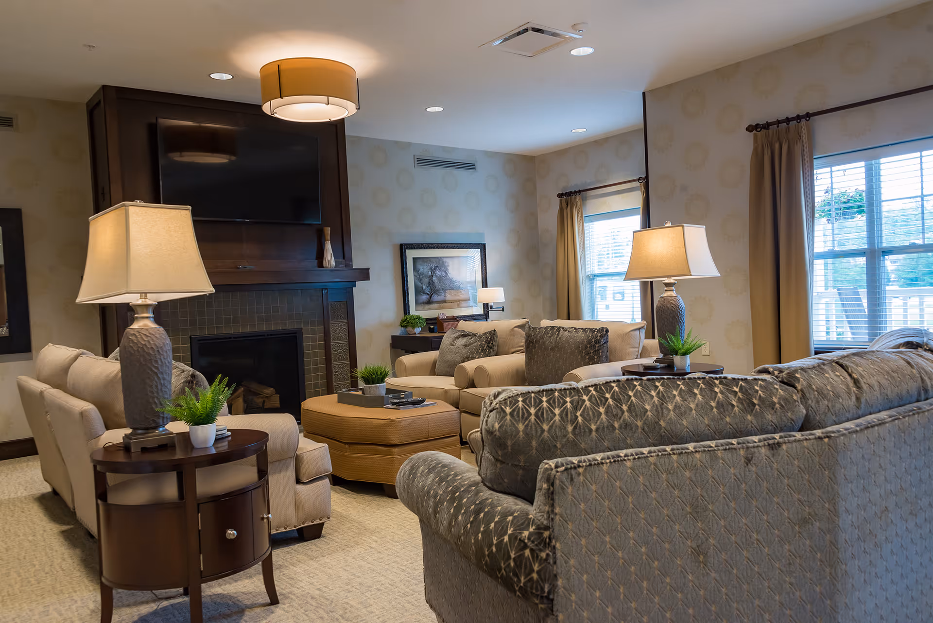 A cozy living room with multiple sofas and armchairs arranged around a central ottoman. The room features a fireplace with a mounted flat-screen TV above it, two large windows with beige curtains, several table lamps, and decorative plants on side tables. The walls have a subtle patterned wallpaper, and a framed picture hangs on one wall.