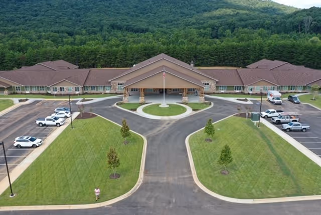 Aerial view of Laurel Lodge Assisted Living & Memory Care facility showing a large single-story building with a covered entrance, surrounded by parking lots and green lawns with small trees. In the background, there is a forested hillside.