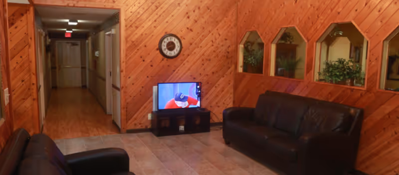 A cozy interior common area with wood-paneled walls and tiled floor. The room features two black leather sofas facing a small TV on a stand. There is a clock on the wall above the TV, and three windows with wooden frames looking into another room with plants visible. A hallway extends from the left side of the room.