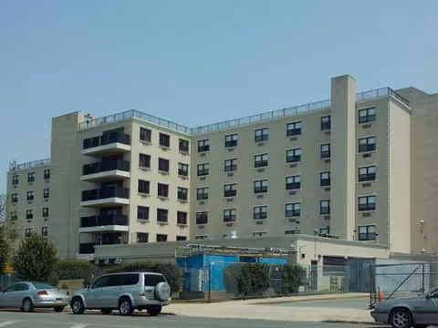 Multi-story beige senior living facility with balconies, a parking lot, and blue construction fencing.
