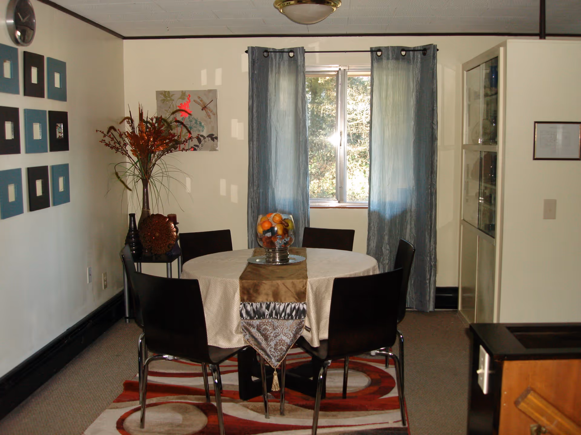 A dining room with a round table covered by a beige tablecloth and a decorative runner. Six dark wooden chairs surround the table. A glass bowl filled with artificial fruit sits in the center. Behind the table is a window with blue curtains. To the left, there is a small table with decorative vases and a wall adorned with square art pieces. The floor has a patterned rug under the table.