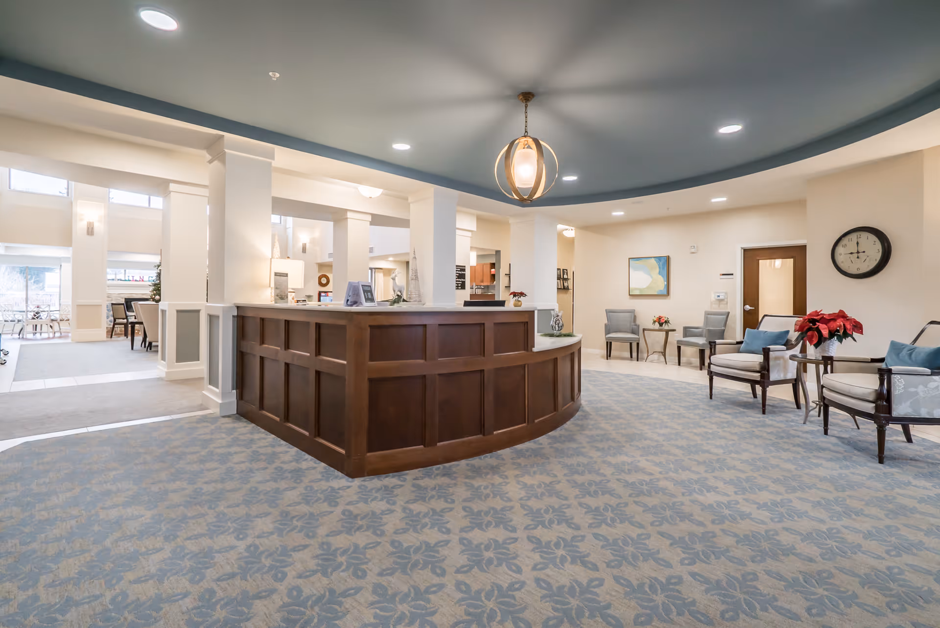 A spacious and well-lit reception area in a senior living facility featuring a large wooden reception desk in the center, patterned carpet flooring, several seating areas with chairs and small tables, a wall clock, and modern ceiling lights including a central hanging light fixture.