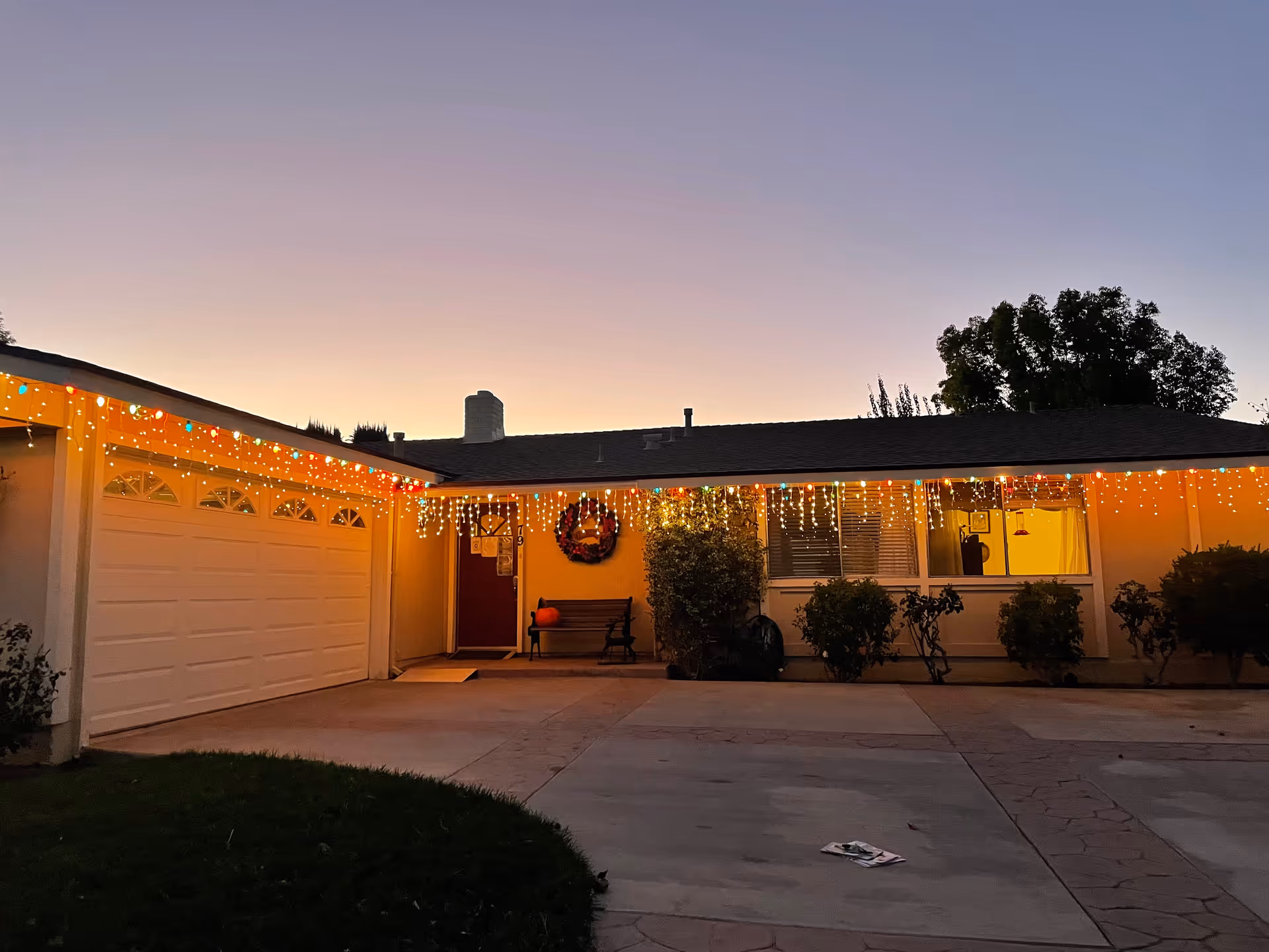 Exterior view of a single-story house at dusk with holiday string lights hanging along the roofline. The house has a garage on the left, a red front door decorated with a wreath, a bench with a pumpkin, and several bushes in front of the windows. The sky is clear with a gradient from light to dark.