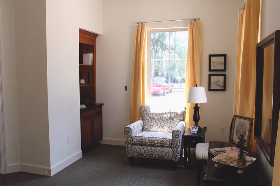 A cozy corner of a living room with a patterned armchair placed in front of a large window with yellow curtains. Next to the chair is a small black side table with a lamp and a decorative bowl. On the right side, there is a dark wooden dresser with framed pictures and a decorative cross. A built-in wooden cabinet is visible on the left side of the image.