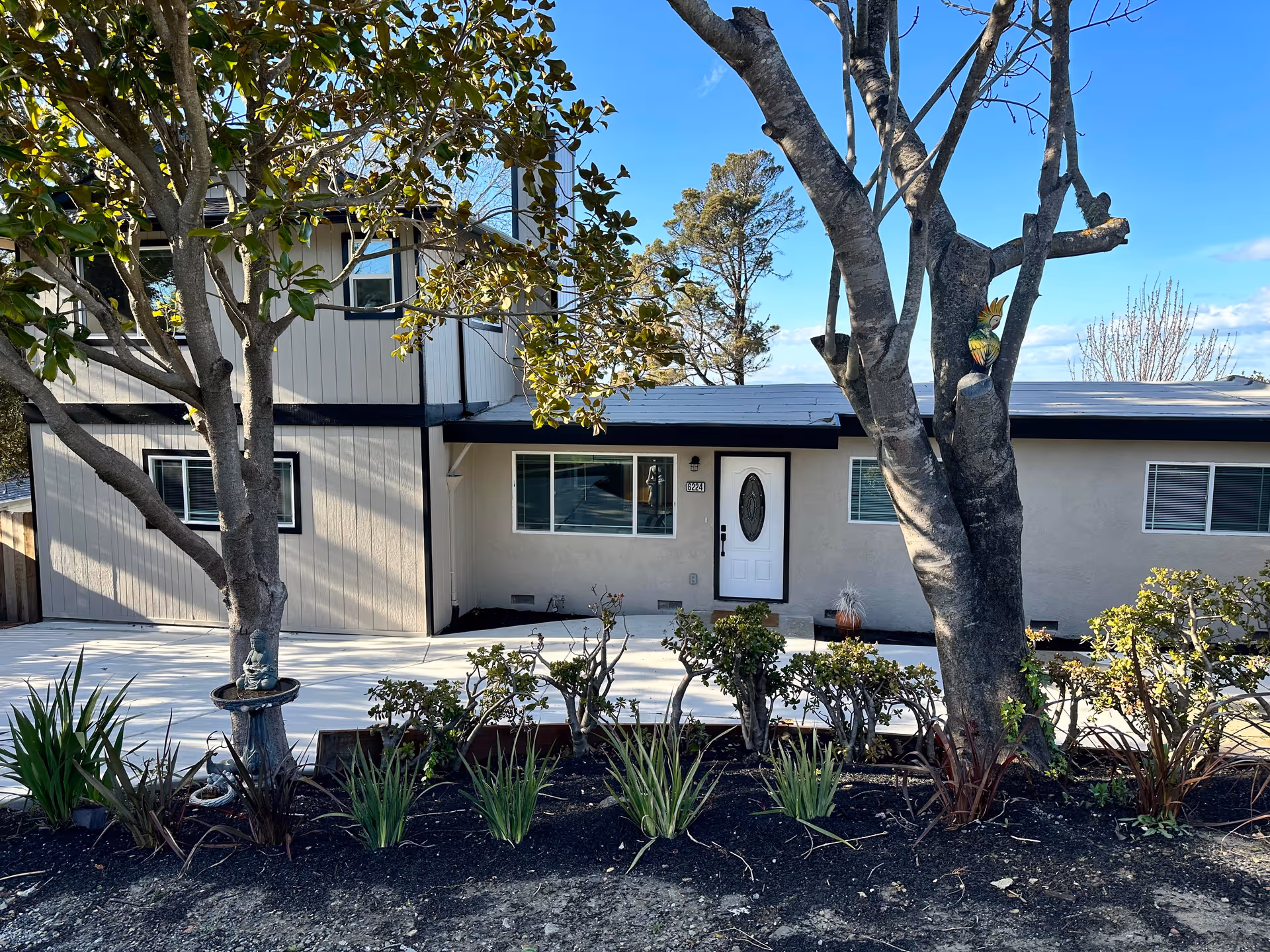 Exterior view of a two-story residential building with beige walls and black trim. There are two large trees and various plants in front of the building, along with a concrete pathway leading to a white front door with an oval glass panel. The sky is clear and blue.