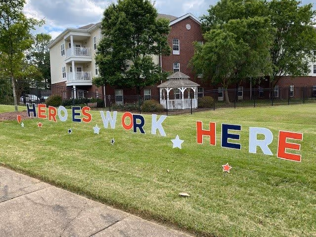 Outdoor grassy area in front of a multi-story senior living building with balconies and a white gazebo. Large colorful letters on the grass spell out 'HEROES WORK HERE' with star decorations around the letters.