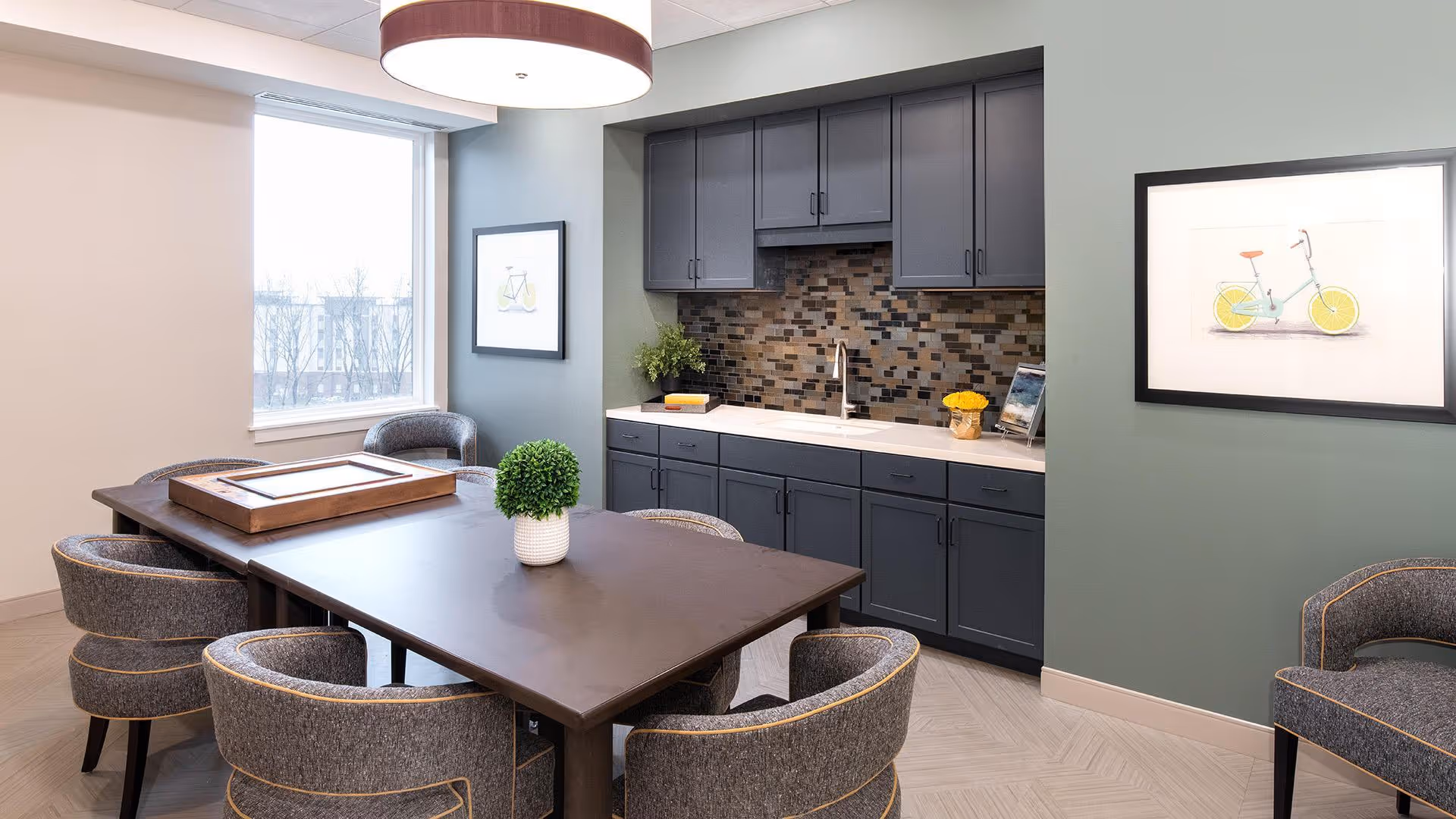 A modern dining area with a dark wooden table surrounded by six upholstered chairs with gray fabric and yellow piping. Behind the table is a kitchenette with dark gray cabinets, a white countertop, a mosaic tile backsplash, and a sink. The walls are painted in light and muted green tones, decorated with framed pictures of bicycles. A large window lets in natural light, and a round ceiling light fixture hangs above the table. A small potted plant is placed on the table.