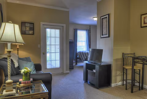 Interior view of a living area in Dalton Square facility featuring a sofa with cushions and a lamp on a side table, a glass door leading outside, a TV on a stand, and a small dining area with two chairs and a table. In the background, a bedroom with a bed and window is visible.