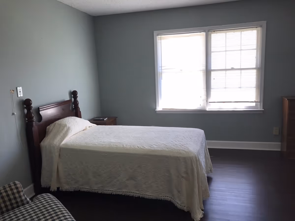 A simple bedroom with a single bed covered in a white bedspread, a wooden headboard, a small wooden nightstand with a book on it, a window with blinds letting in natural light, and a checkered armchair partially visible in the foreground.