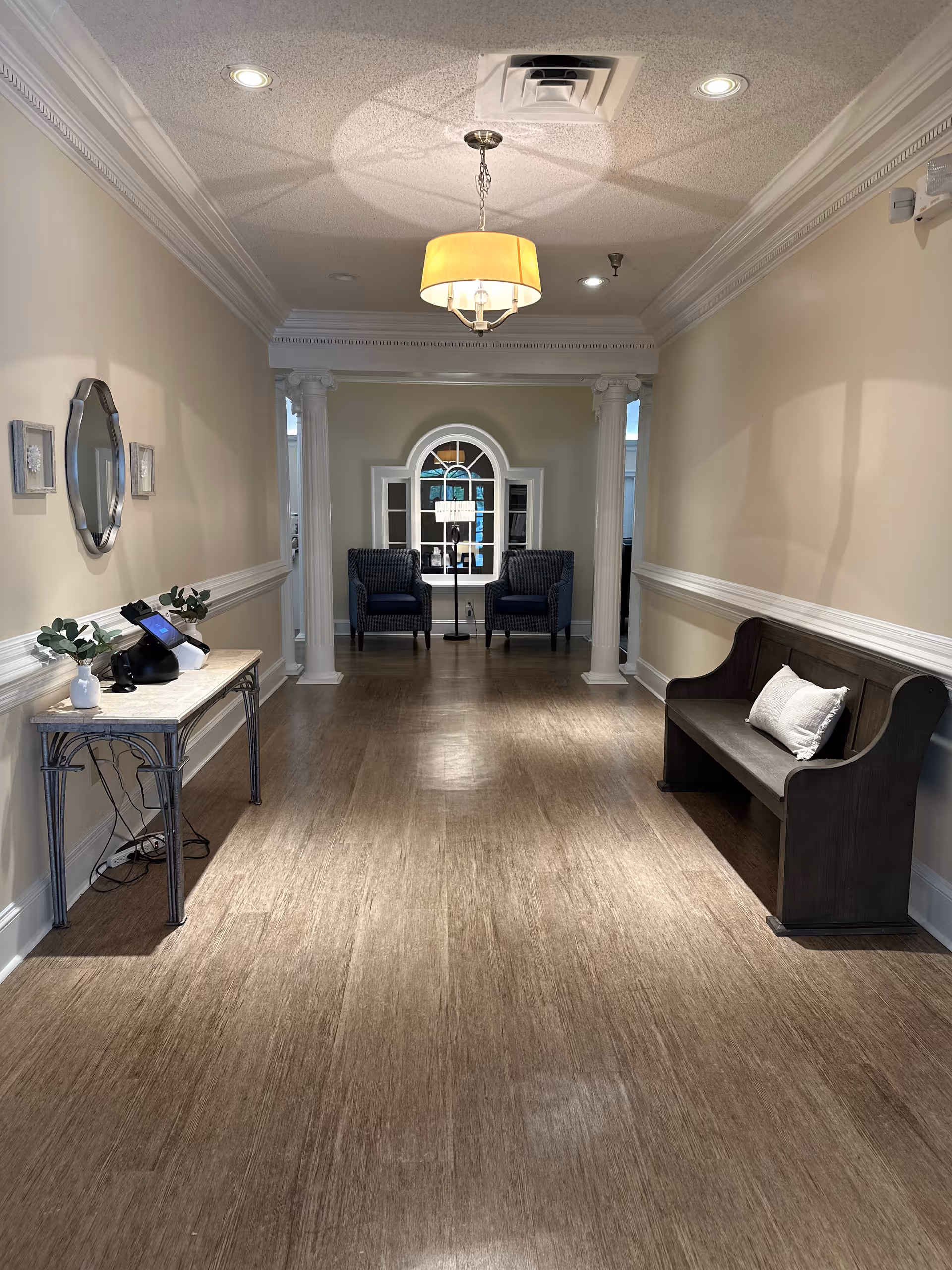 A hallway in a senior living facility with wooden flooring, cream-colored walls, and white crown molding. On the left side, there is a small table with a phone and decorative plants, and a mirror above it. On the right side, there is a dark wooden bench with a white pillow. At the end of the hallway, two blue armchairs are positioned in front of a window with an arched top, flanked by two white columns. A ceiling light fixture hangs in the center of the hallway.