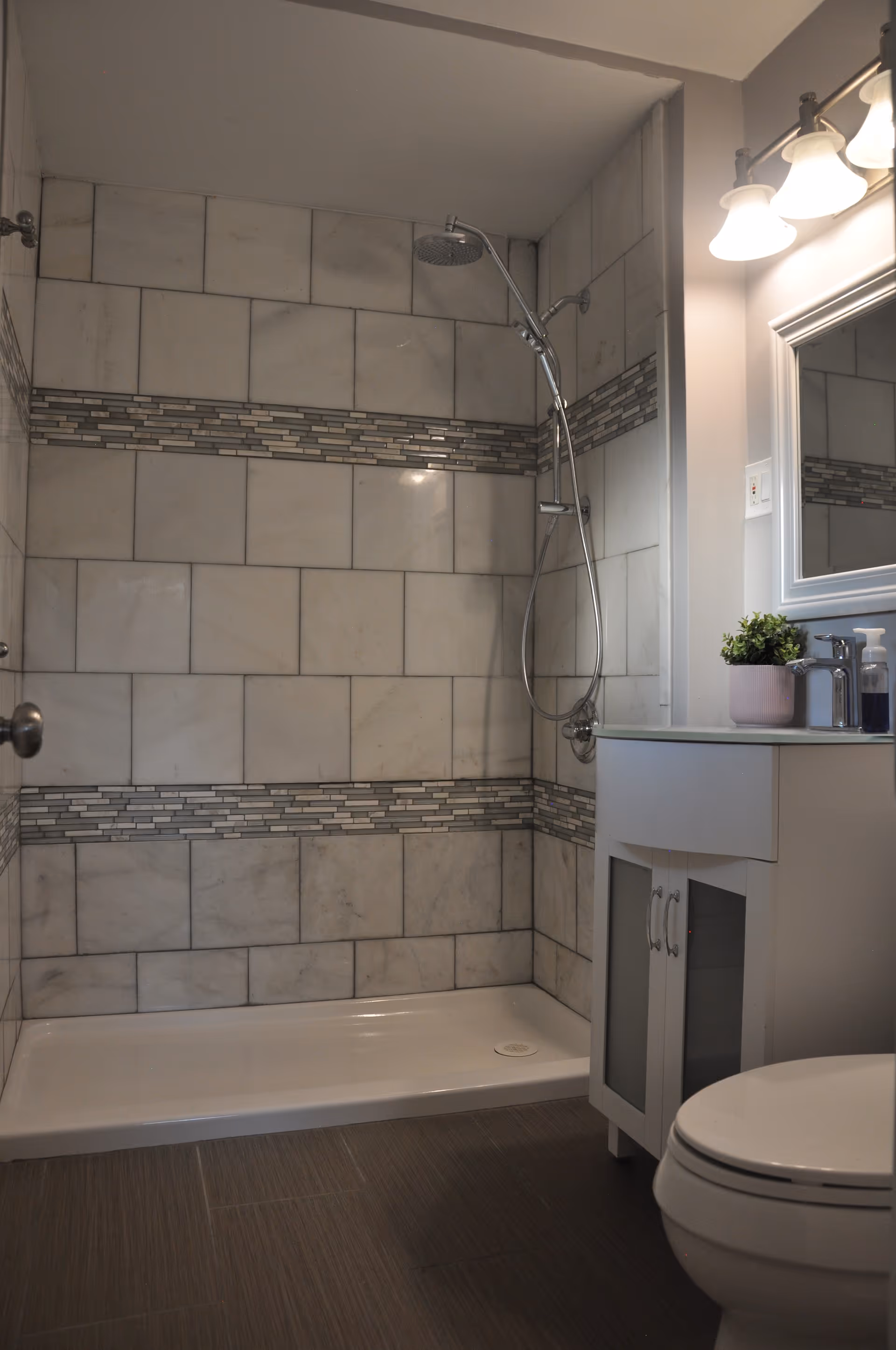 Small modern bathroom with a tiled walk-in shower featuring mosaic accent bands, a chrome showerhead, a white vanity with mirror, and a toilet.