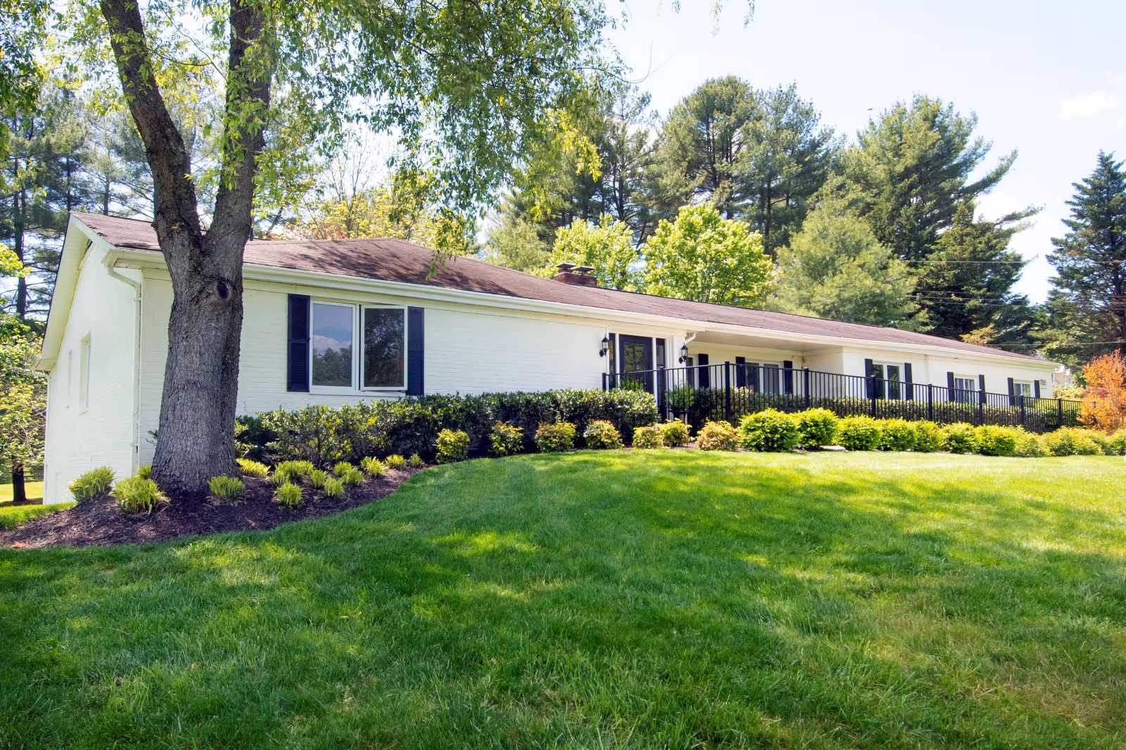 Single-story white brick building with a brown roof surrounded by green grass, bushes, and tall trees under a clear sky.