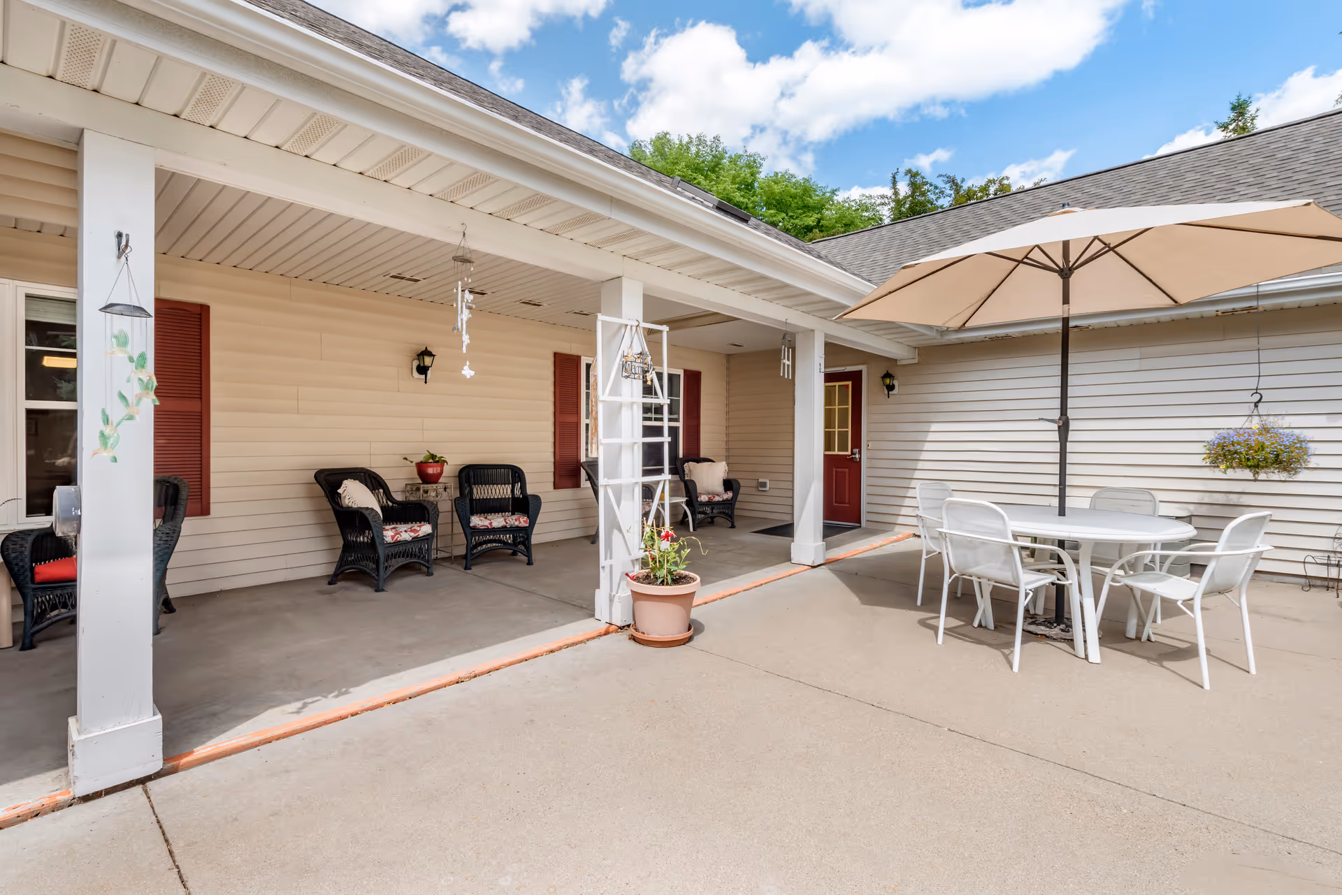 Outdoor patio area with a covered seating section featuring black wicker chairs with cushions and a small table. There is also a white round table with six white chairs under a large beige umbrella. The building exterior is beige with red shutters and a red door. Potted plants and hanging decorations are visible.