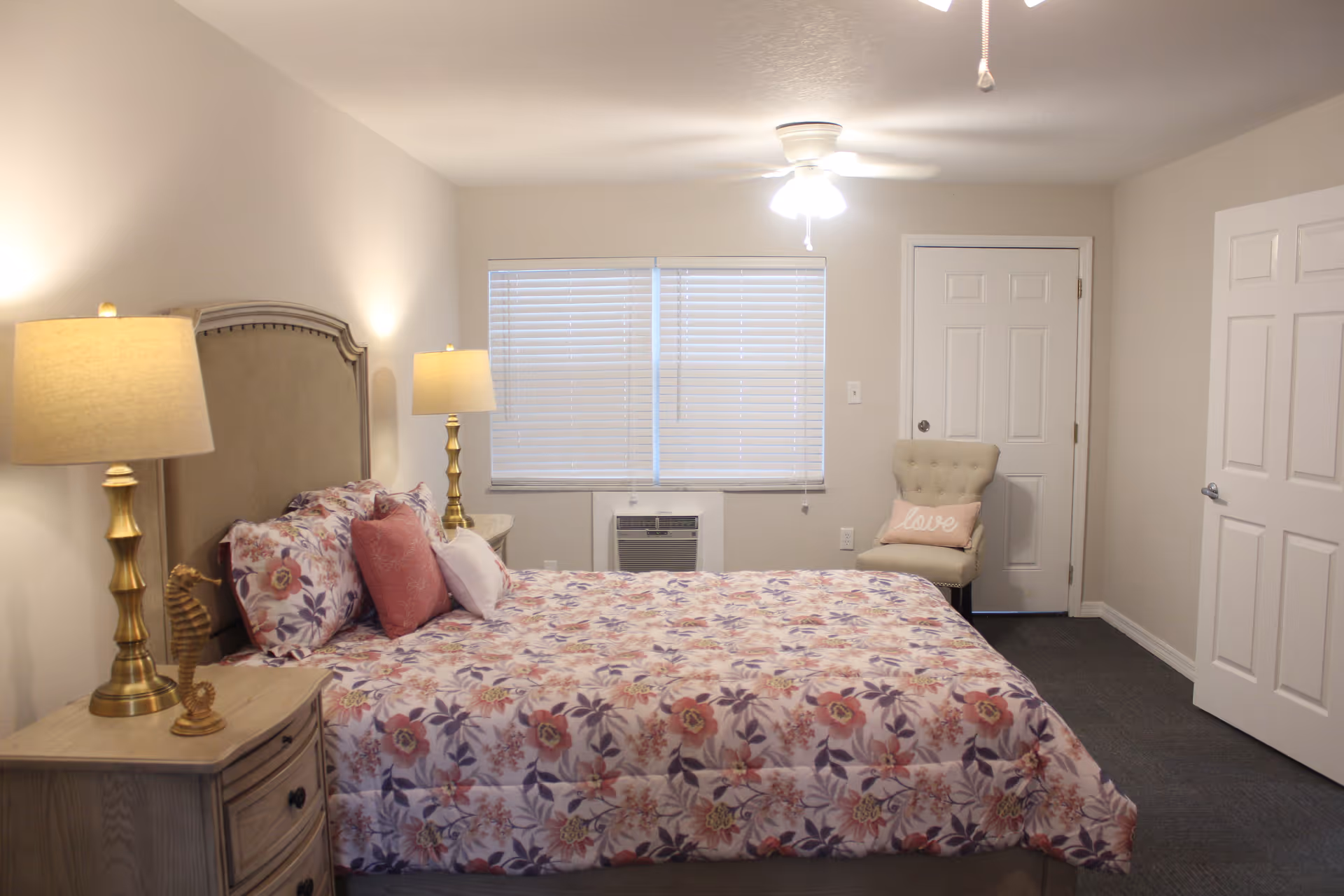 A cozy bedroom with a floral patterned bedspread on a bed with a cushioned headboard. Two bedside tables each have a brass lamp with a beige shade. A beige upholstered chair with a pink pillow that says 'love' is placed near a closed door. The room has a window with closed blinds and an air conditioning unit below it. The walls are painted light beige and the floor is carpeted in dark gray.