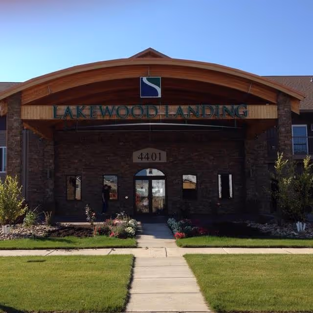 Front entrance of the Lakewood Landing building with an arched wooden canopy, stone facade, address plaque reading 4401, and a sidewalk through a lawn to the doors.