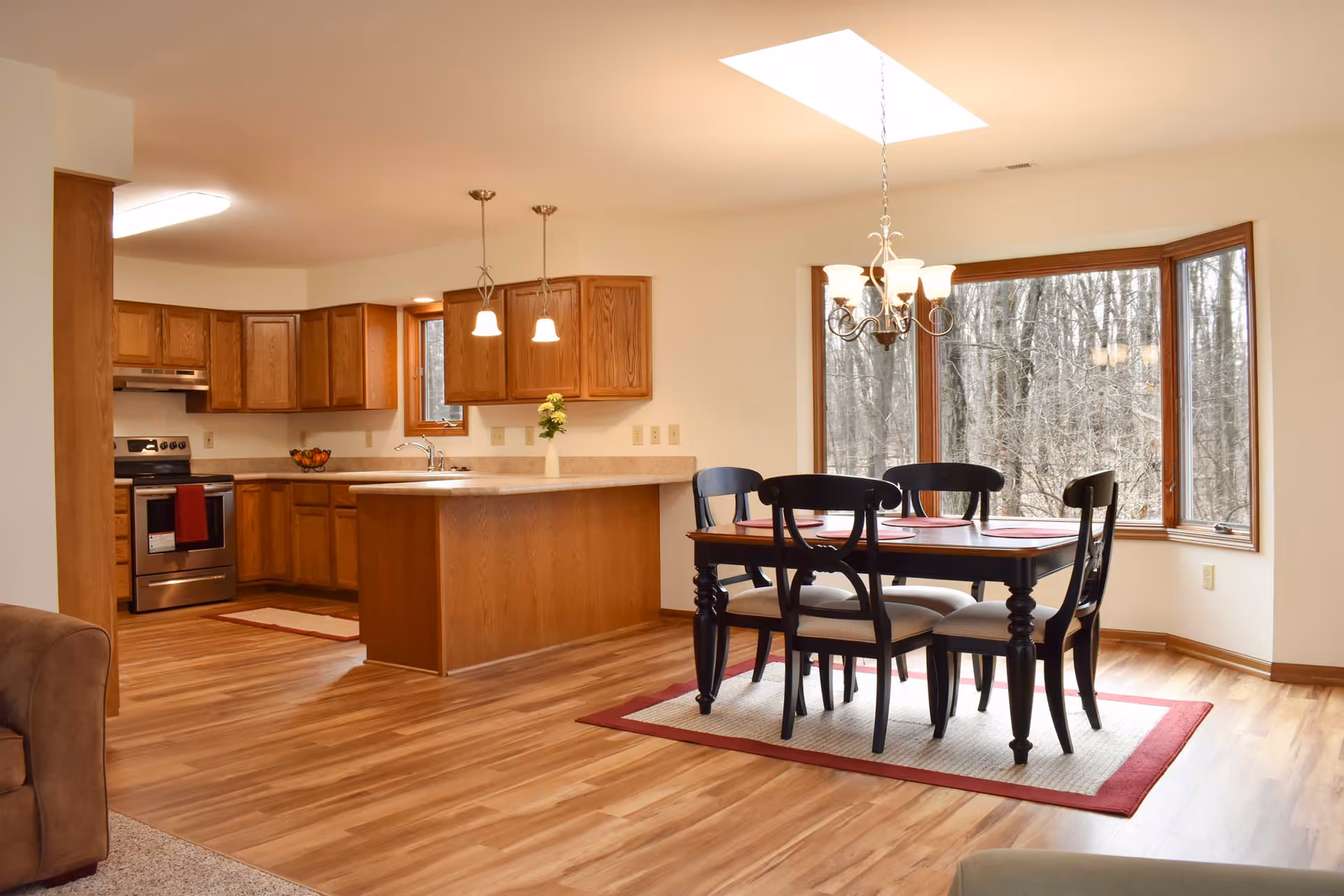 Open-concept kitchen and dining area with a wooden table and chairs on a rug beside a large bay window and a kitchen island with pendant lights.