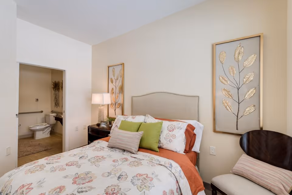 Cozy bedroom with a floral bedspread, nightstand and lamp, and an open doorway revealing a bathroom.