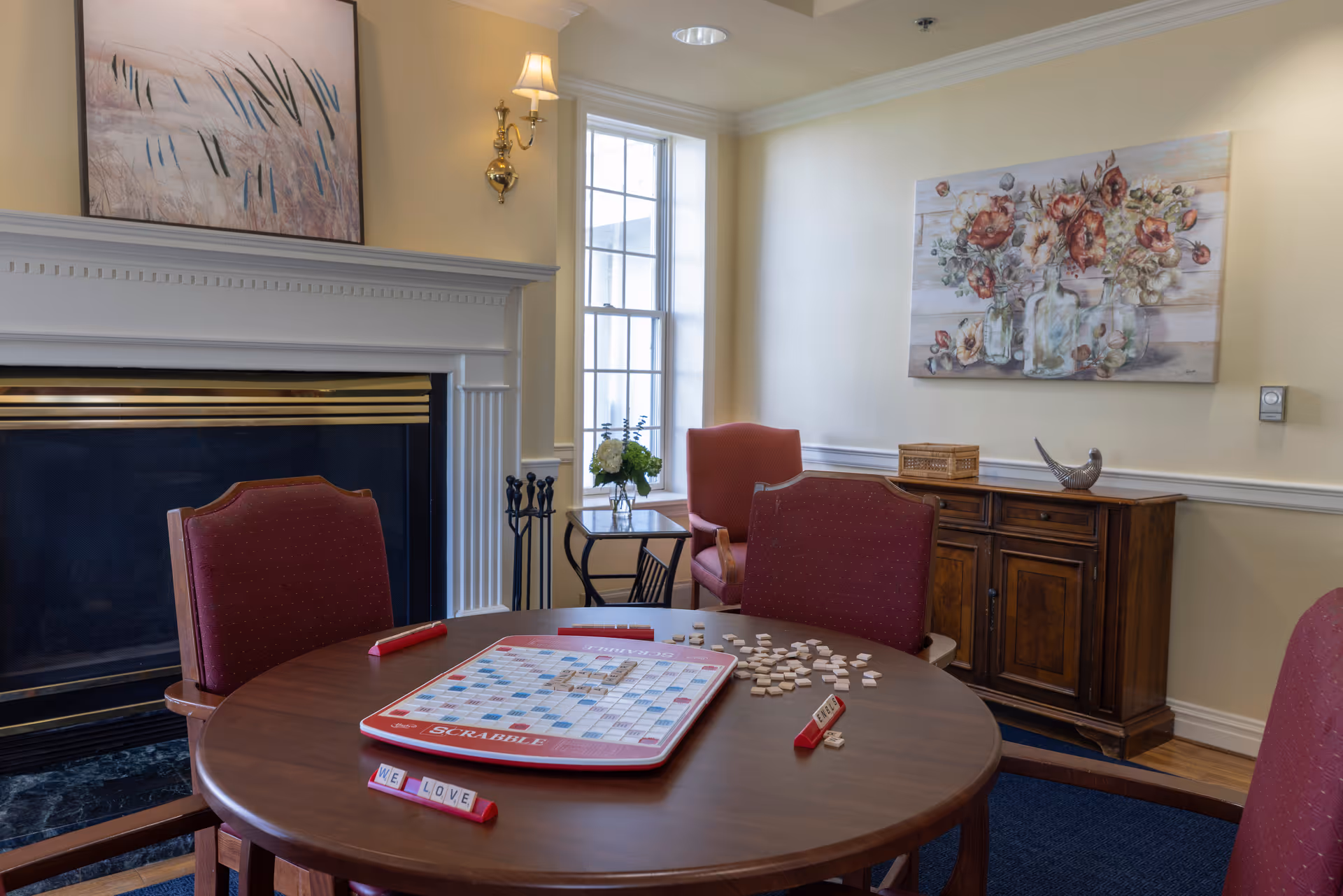 A cozy room with a round wooden table set for a game of Scrabble, surrounded by four red upholstered chairs. The table has a Scrabble board with tiles and racks, one rack displaying the words 'WE LOVE'. Behind the table is a fireplace with a painting above it, a window letting in natural light, and a wooden cabinet with a floral painting and decorative items on top.