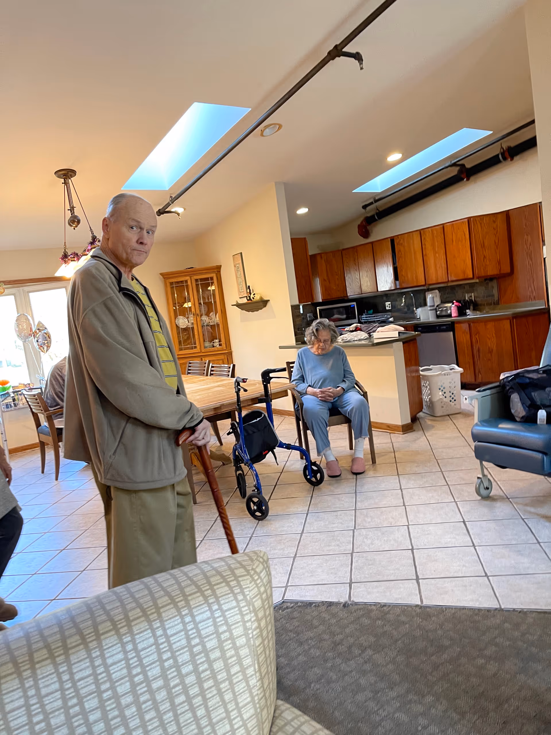 An elderly man with a cane standing in a living area with tiled floors and a kitchen in the background. An elderly woman is seated on a chair near a walker. The room has wooden cabinets, a dining table with chairs, and skylights on the ceiling.