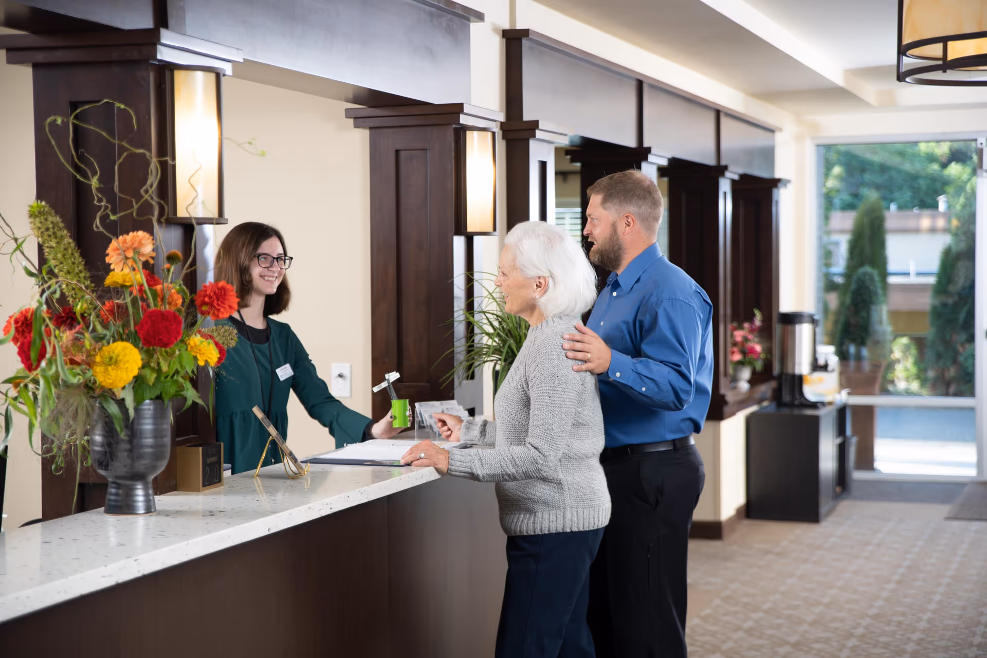 An elderly woman and a middle-aged man standing at a reception desk inside a senior living facility, interacting with a smiling female receptionist. The reception area features a large flower arrangement on the counter, dark wood paneling, and a bright, welcoming atmosphere with natural light coming through a glass door in the background.