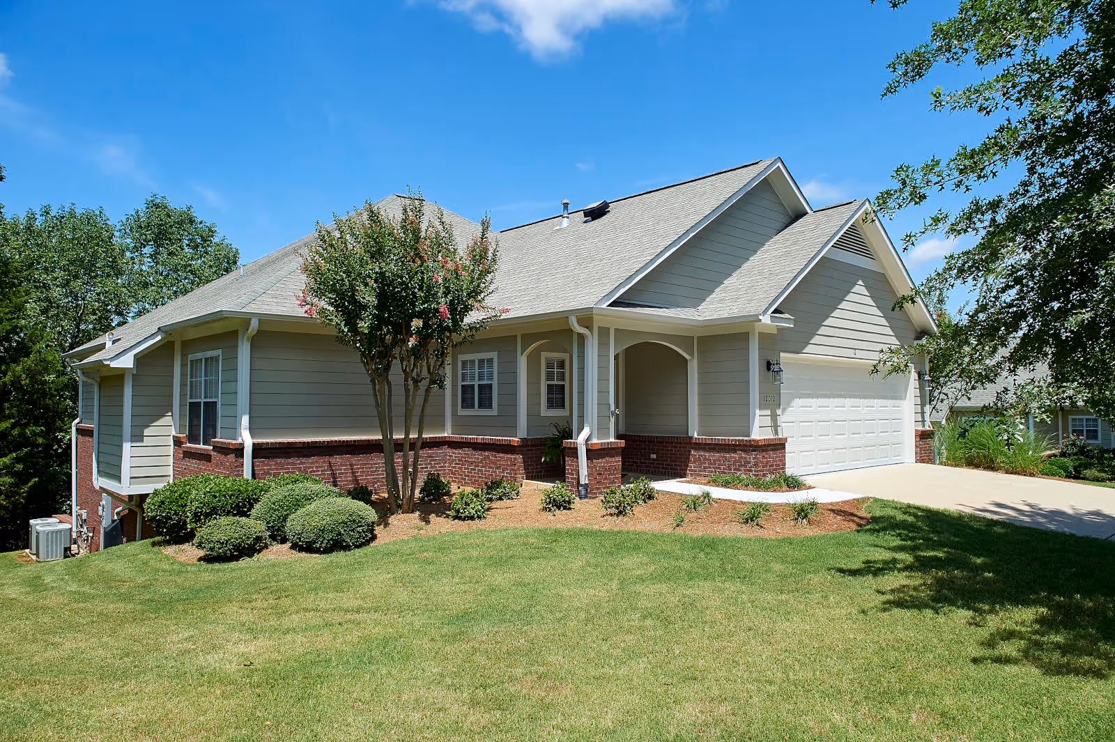 Exterior view of a single-story residential building with beige siding and red brick accents, a two-car garage, a well-maintained lawn, shrubs, and a tree in front under a clear blue sky.