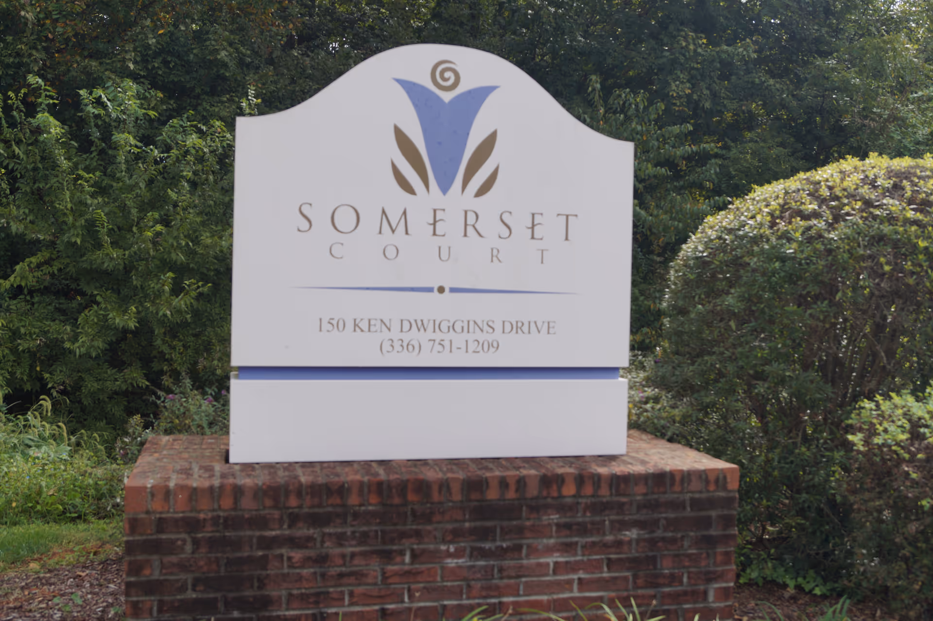 White entrance sign reading "Somerset Court" with the address on a brick base surrounded by shrubs and trees.