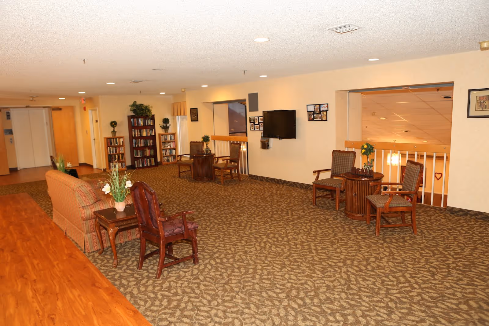 A spacious senior living facility common area with patterned carpet and beige walls. The room features several wooden chairs with cushions, small round tables with flower arrangements, a couch, and bookshelves filled with books. A flat-screen TV is mounted on the wall, and there are framed pictures hanging nearby. The area is well-lit with ceiling lights and has a wooden floor section on one side.