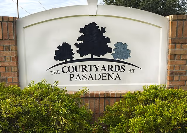 A white sign with black silhouettes of three trees and the text 'The Courtyards at Pasadena' mounted on a brick base with green bushes in front.