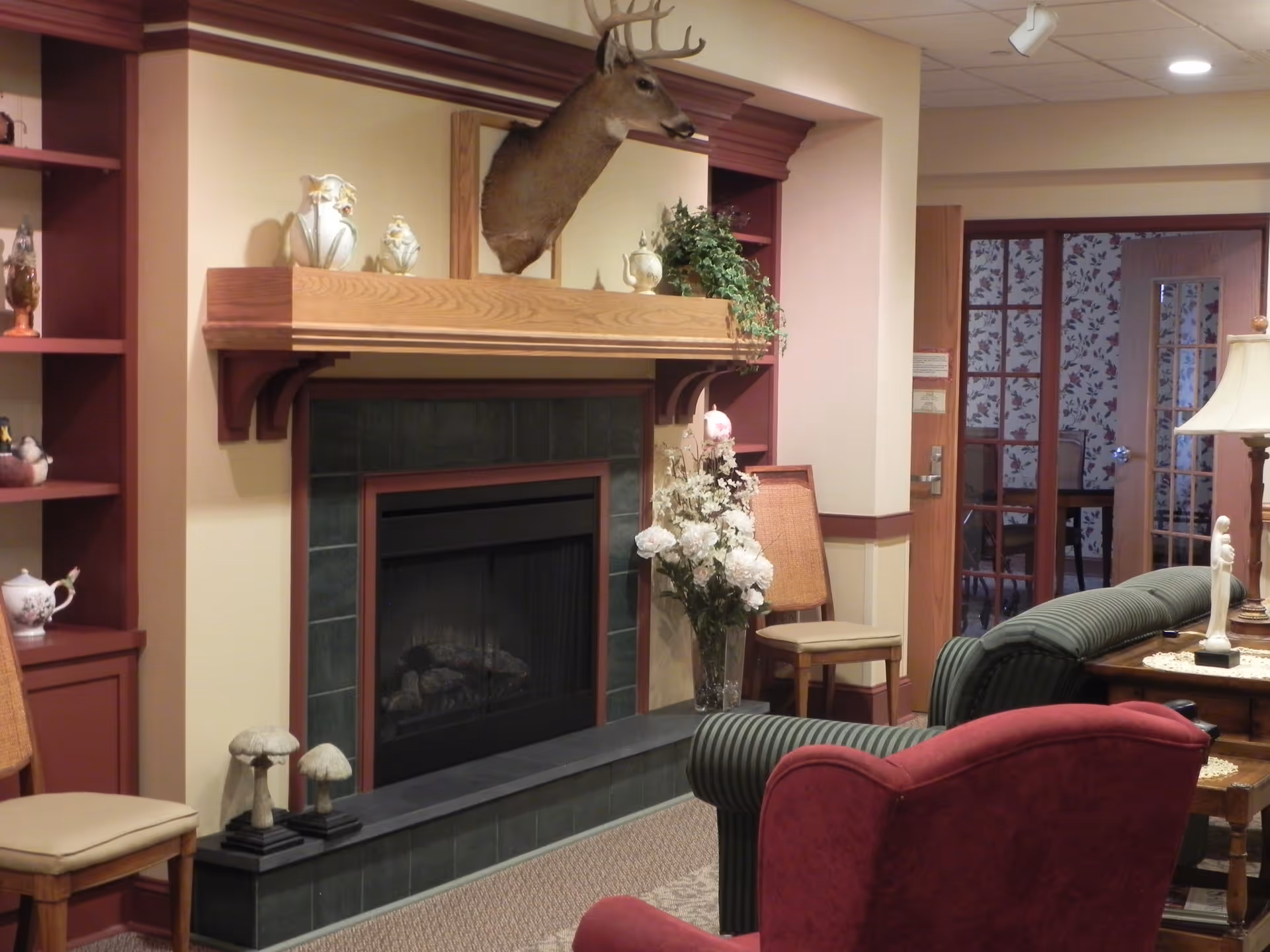 A cozy living room area in a nursing home featuring a fireplace with a mounted deer head above it. The fireplace is surrounded by dark green tiles and a wooden mantel decorated with ceramic figurines and a plant. There are upholstered chairs, a side table with a lamp, and floral arrangements near the fireplace. In the background, there is a doorway with glass-paneled doors leading to another room with floral wallpaper.