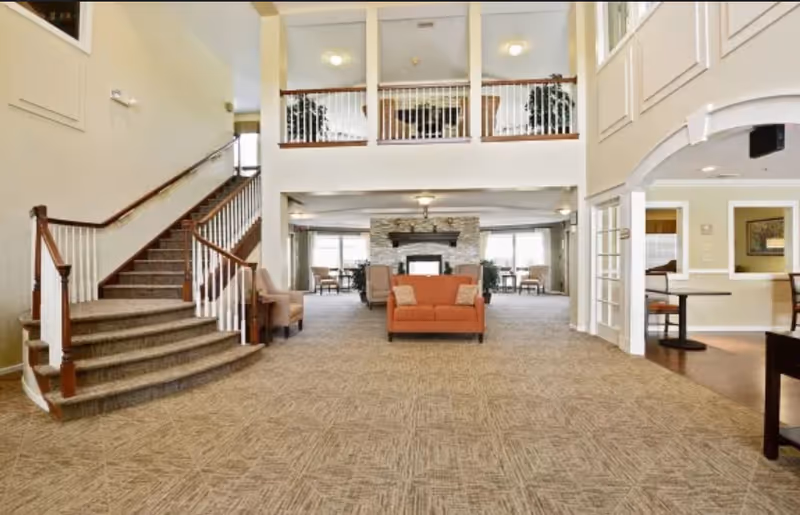 Spacious senior living facility common area with a carpeted floor, a staircase with wooden handrails on the left, an orange loveseat in the center, and a stone fireplace in the background. The area has high ceilings with a second-floor balcony overlooking the space and several seating areas with chairs and tables.