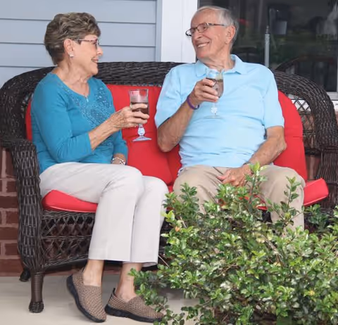 An elderly man and woman sitting on a wicker loveseat with red cushions on a porch, smiling and holding glasses of a beverage, with green plants in the foreground.