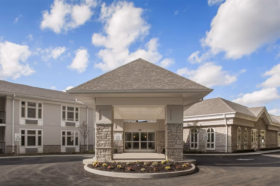 Front entrance of a senior living facility with a covered drop-off area supported by stone pillars, surrounded by a circular flower bed. The building has beige walls, multiple windows, and a gray shingled roof under a partly cloudy blue sky.