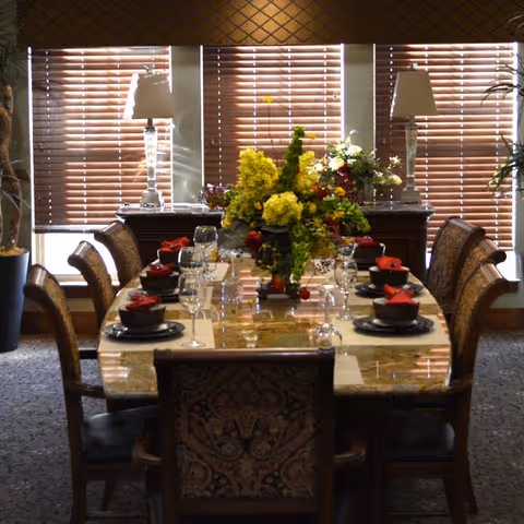 A formal dining room with a long rectangular table set for eight people. The table has a polished stone surface and is decorated with a large floral centerpiece featuring yellow and green flowers. Each place setting includes a black bowl, a plate, a wine glass, and a red folded napkin. The room has patterned upholstered chairs, two table lamps on a sideboard against the window, and wooden blinds covering the windows in the background.