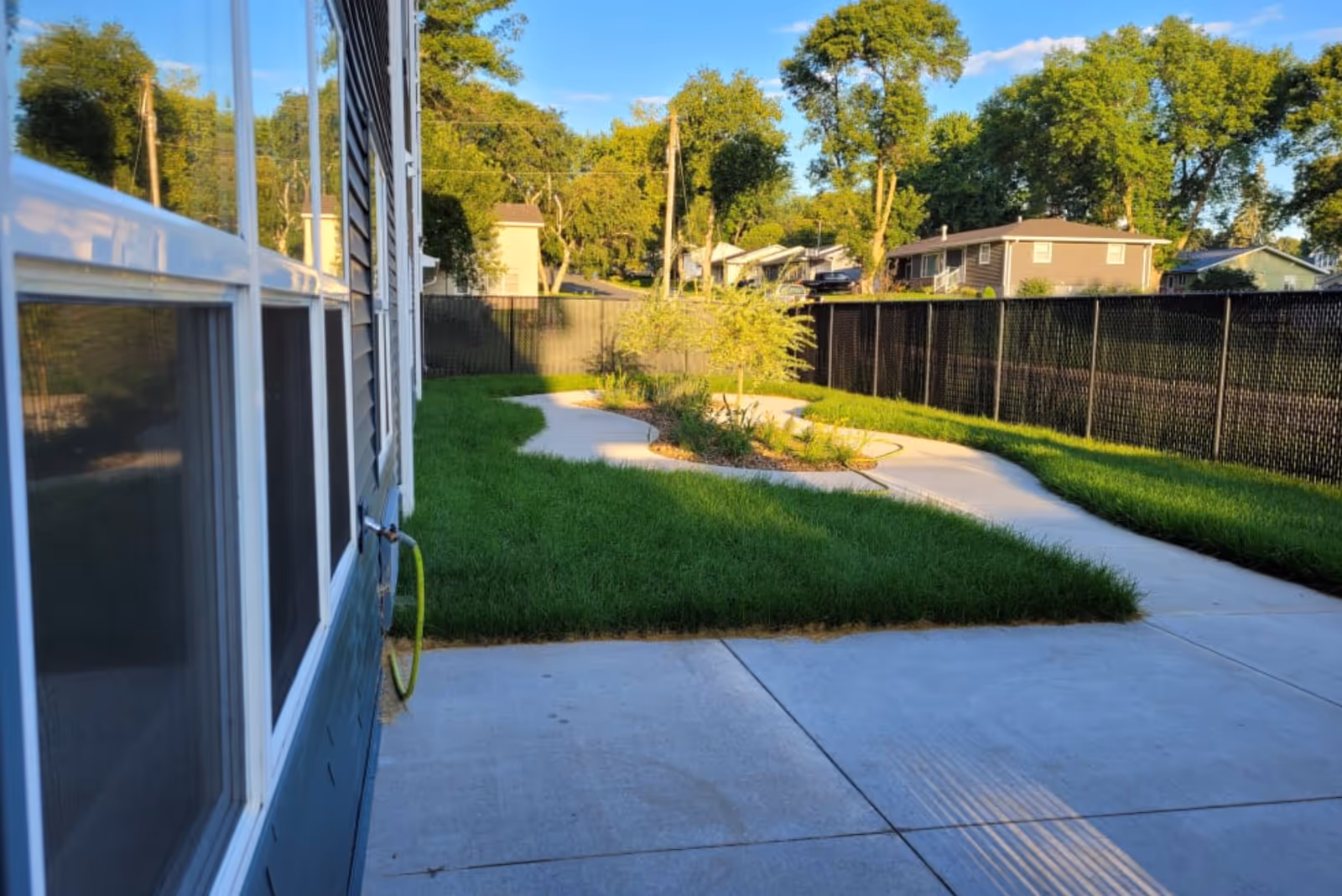 Outdoor view of a fenced yard at Goodhue Living facility with a concrete patio, green grass, a curved concrete pathway, and a small landscaped area with plants and a young tree. Residential houses and tall trees are visible in the background under a clear blue sky.
