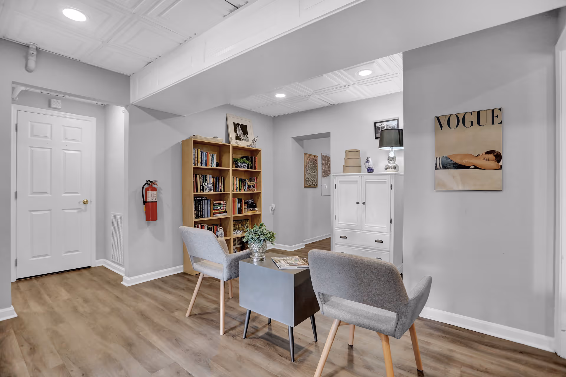 A cozy interior room with light gray walls and wood flooring featuring two gray upholstered chairs around a small square table with a plant and magazines on it. There is a wooden bookshelf filled with books and decorative items against one wall, a white cabinet with a lamp and decorative boxes on top, and a framed Vogue magazine cover hanging on the wall. A fire extinguisher is mounted on the wall near a white door.