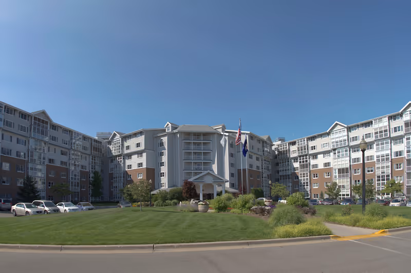 Exterior view of a large multi-story senior living facility building with a well-maintained lawn, landscaped garden, and two flagpoles displaying the American flag and another flag. Several cars are parked along the driveway under a clear blue sky.