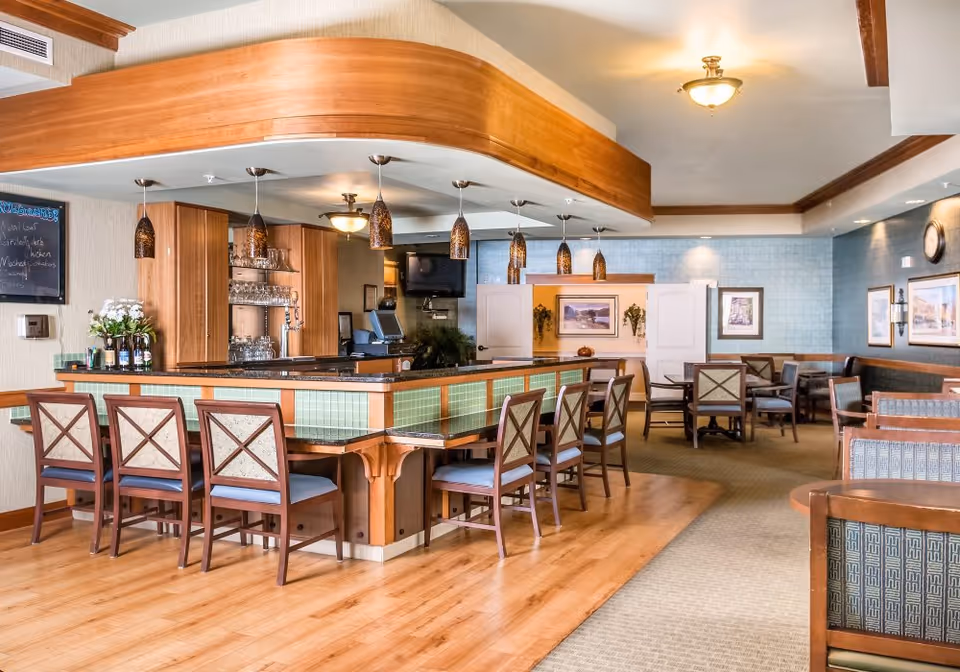 A spacious dining area with a wooden bar counter surrounded by chairs. The bar has hanging pendant lights and shelves with glasses. The room features wooden flooring near the bar and carpeted flooring in the seating area with multiple tables and chairs. The walls are decorated with framed pictures and a clock, and the ceiling has recessed lighting and crown molding.