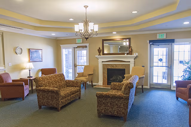 A cozy senior living facility common area with patterned armchairs and sofas arranged around a fireplace. The room has a chandelier, recessed lighting, a wall clock, framed artwork, and two exit doors with glass panels letting in natural light.