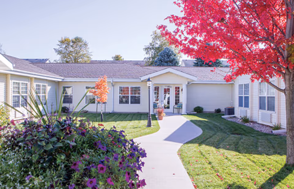 Exterior view of Rolling Meadows Senior Living facility showing a single-story building with beige siding, multiple windows, and a central entrance with glass doors. A concrete pathway leads to the entrance, surrounded by green grass, colorful flowers, and trees with red and orange autumn leaves under a clear sky.