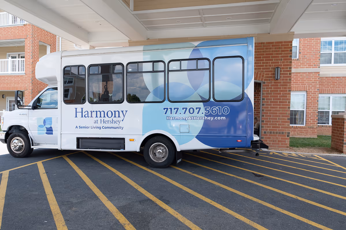 A white and blue shuttle bus parked under a covered entrance of a brick building. The bus has the logo and text 'Harmony at Hershey, A Senior Living Community' along with a phone number and website on its side.