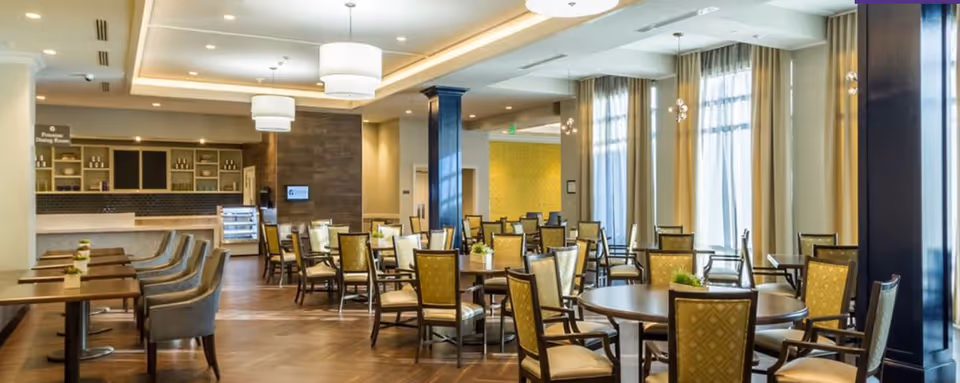 A spacious and well-lit dining room with multiple round and rectangular tables surrounded by cushioned chairs. The room features large windows with sheer curtains allowing natural light to fill the space. The ceiling has modern light fixtures, and there is a counter area in the background with shelves and a display case.