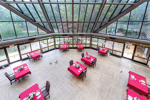 A spacious dining area with large floor-to-ceiling windows and a glass ceiling allowing natural light to fill the room. The room contains several tables covered with red tablecloths, each set with plates, cups, and floral centerpieces. Outside greenery is visible through the windows.