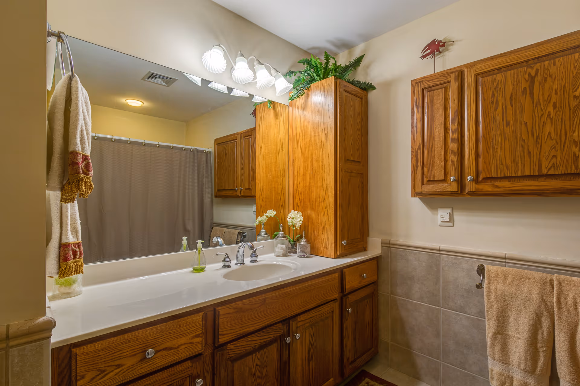 A bathroom with wooden cabinets and a large mirror above a white countertop with a sink. There are decorative plants and containers on the counter, a towel hanging on a ring to the left, and a beige towel hanging on a bar to the right. A shower curtain is visible in the background.