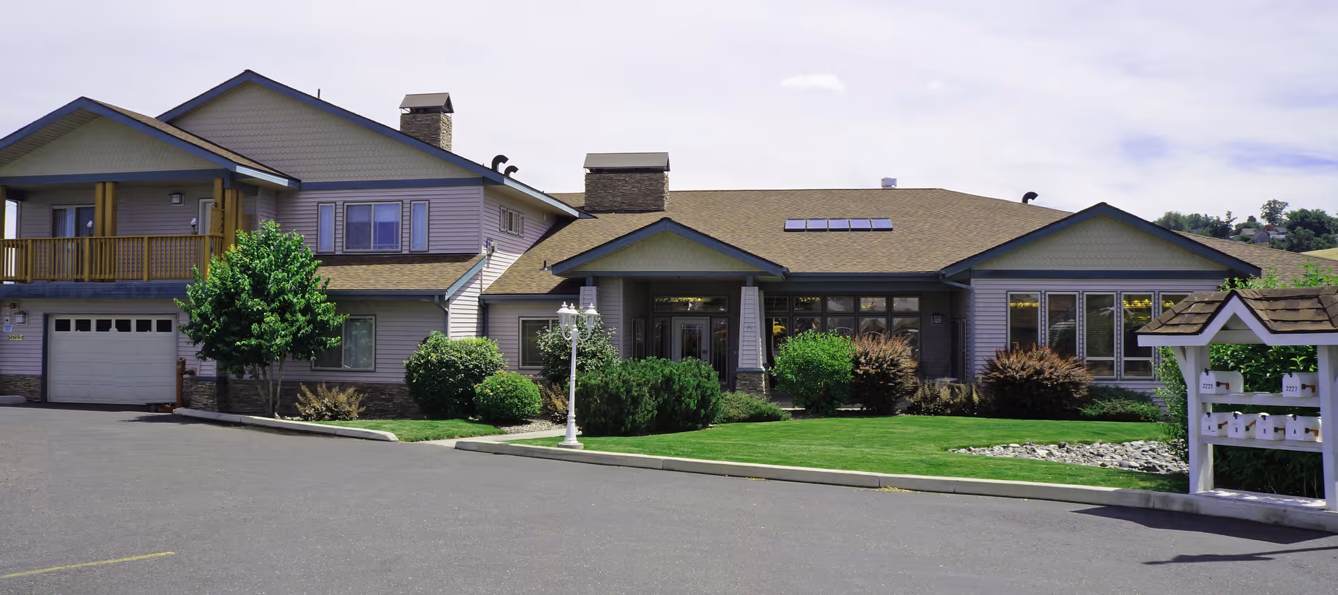 Exterior view of a single-story building with a sloped roof, multiple windows, and a small porch entrance. The building is surrounded by well-maintained green bushes, a tree, and a lawn. There is a paved driveway and a white lamp post near the entrance. On the right side, there is a white mailbox structure with multiple mailboxes.