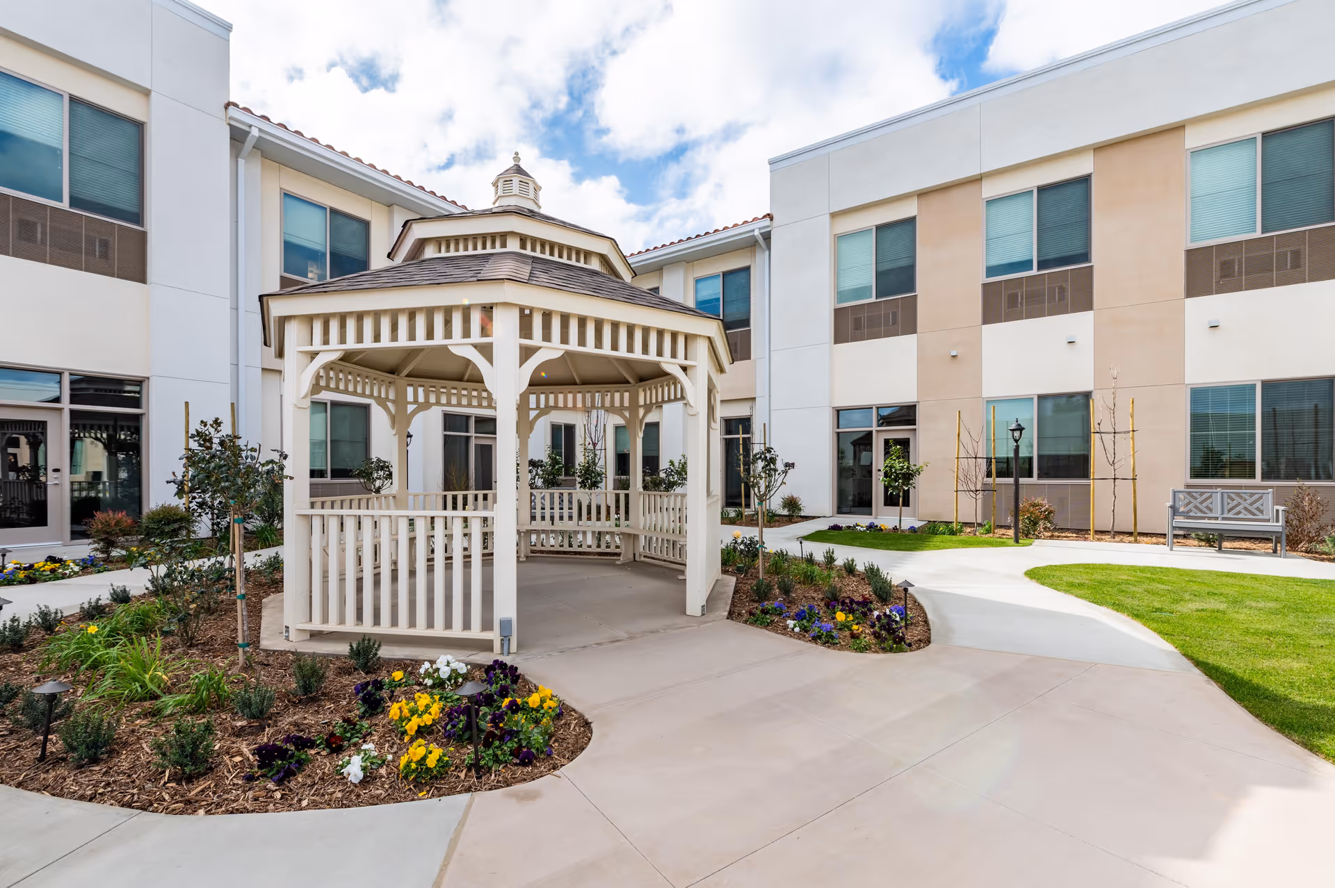 Outdoor courtyard area of a senior living facility featuring a white wooden gazebo surrounded by flower beds and landscaped greenery, with a two-story beige and white building in the background under a partly cloudy sky.